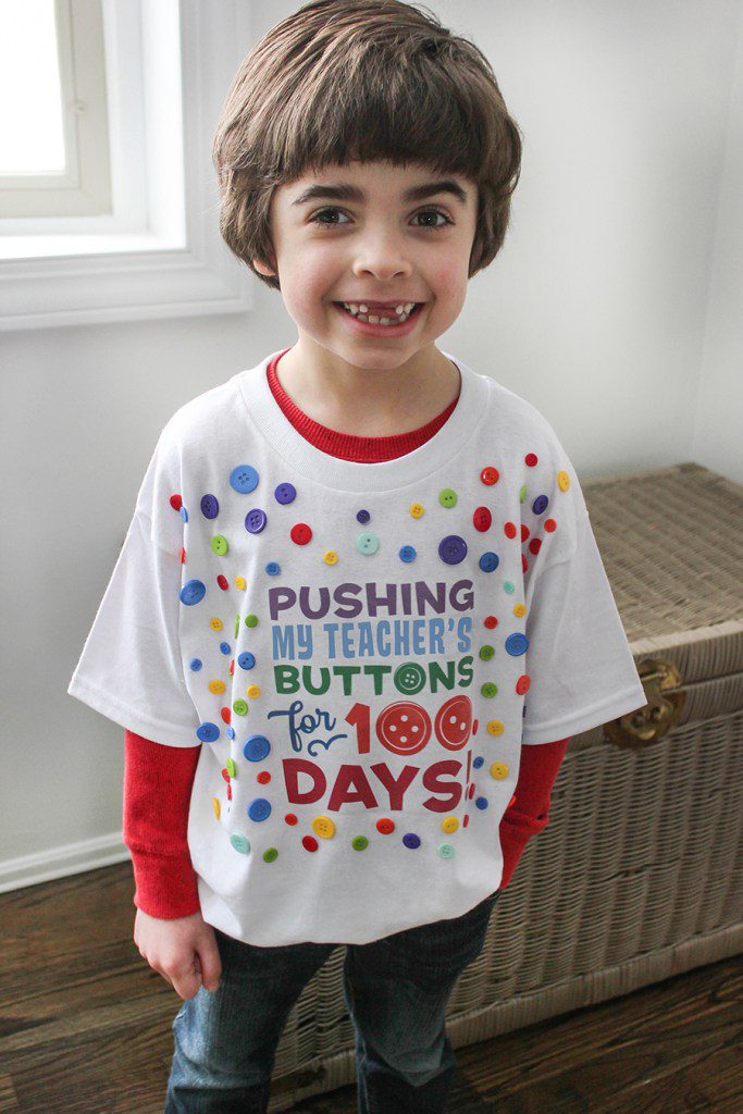 100th day of school shirt ideas include this one. A little boy is wearing a white t-shirt that says Pushing His Teacher's Buttons for 100 Days.