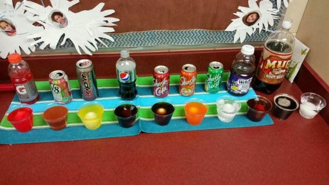 A series of plastic cups filled with varieties of soda, juice, and other liquids, with an egg in each
