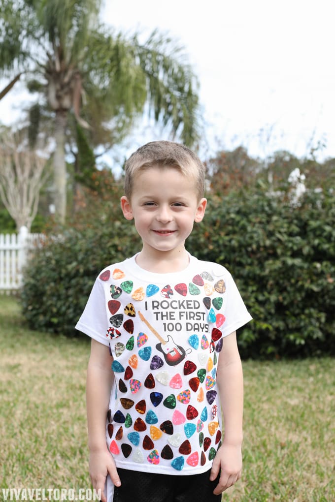A little boy is shown wearing a t-shirt with guitar picks all over it. It says I Rocked the First 100 Days! 