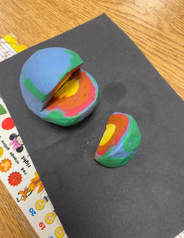 A ball of play doh has a slice taken out of it. It has a yellow center, a brown layer, a red layer, and a blue and green outer shell (first grade science experiments)