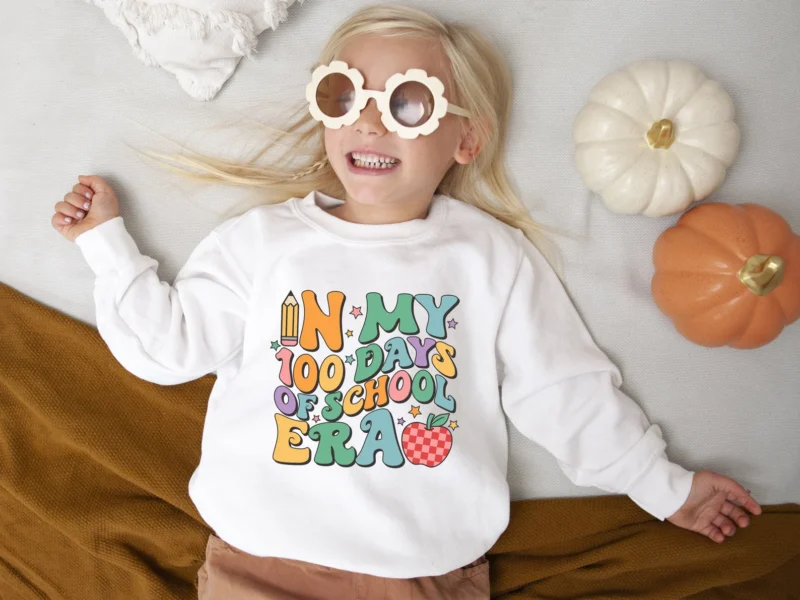 A little girl wearing flower sunglasses is wearing a cream colored sweatshirt that says in my 100 days of school era.