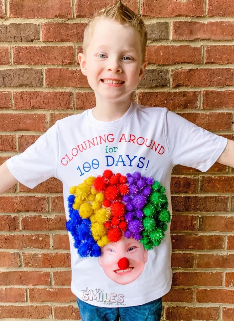 A little boy is wearing a shirt that says clowning around for 100 days! His face is on the shirt with a clown nose and 100 pom poms make up the hair.
